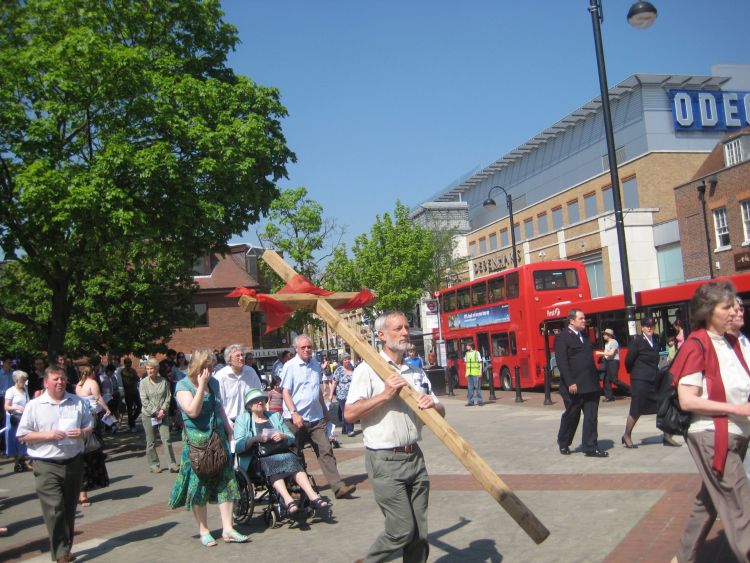 A man walking through Uxbridge town centre carrying a cross with a crowd following behind
